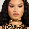 With long, curly hair and a serious expression, an individual wears a leopard print top complemented by the QUIET LUXURY CHOKER. The neutral background highlights their bold fashion choices and makeup, while the choker's metallic faux gold finish enhances their wavy silhouette with an elegant touch.