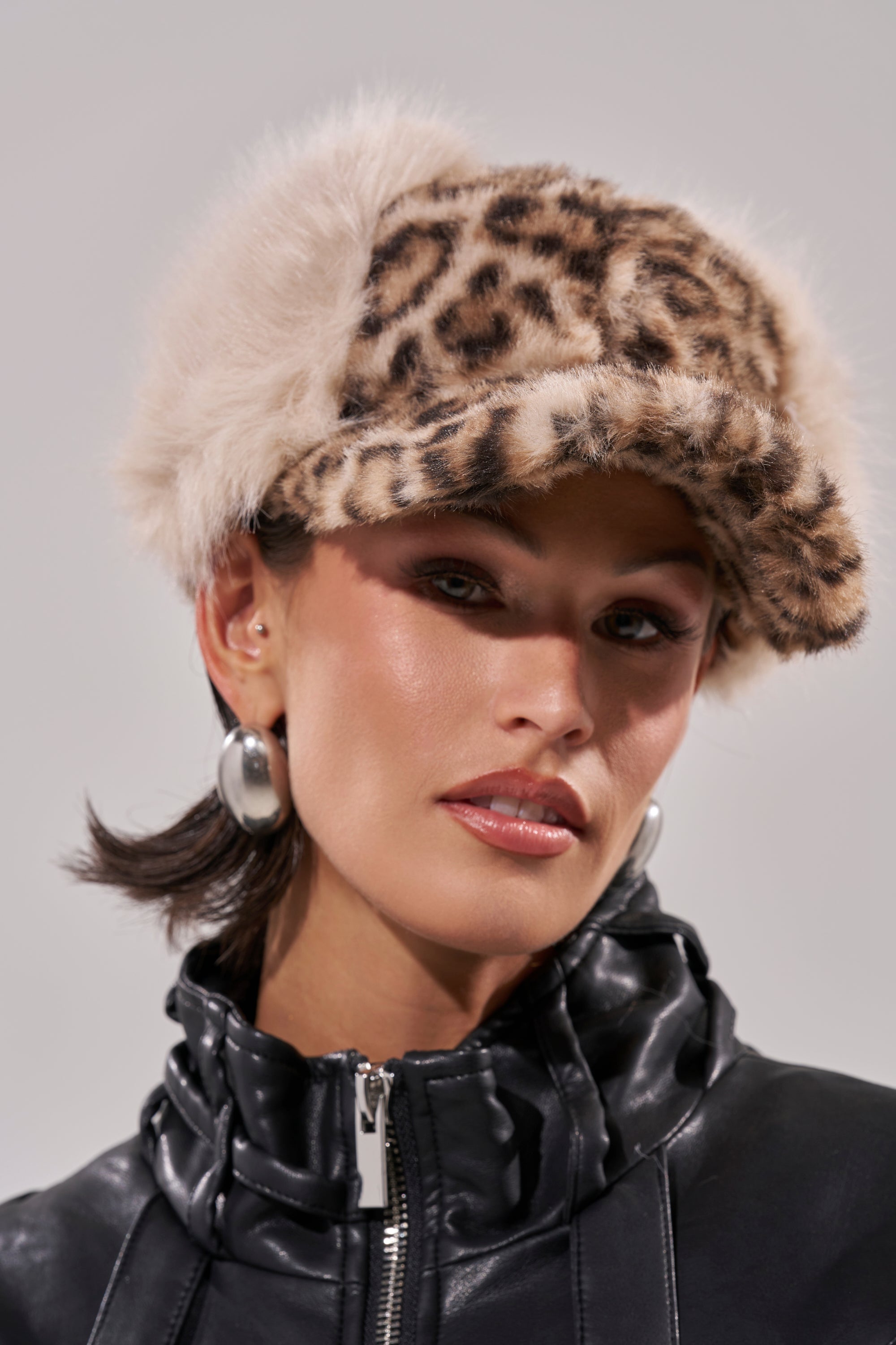 A woman in the ON MY SH** FAUX FUR HAT and silver hoops, wearing a shiny black zip-up jacket, poses against a neutral background. 0