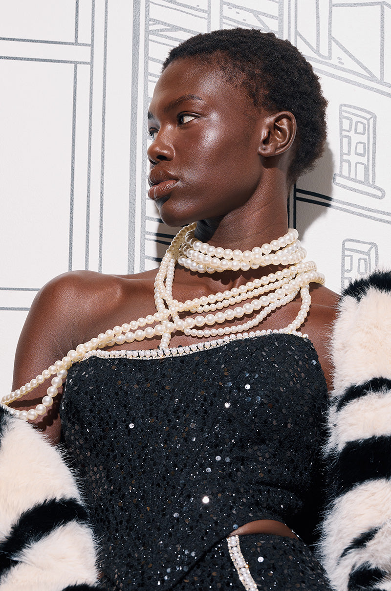 The scene is graced by a person wearing a black sequined dress and the striking PENELOPE STATEMENT PEARL NECKLACE. Their shoulders are elegantly draped with a garment featuring bold black and white stripes, while in the background, a sketch of windows and buildings enhances the layered finish of this sophisticated tableau.