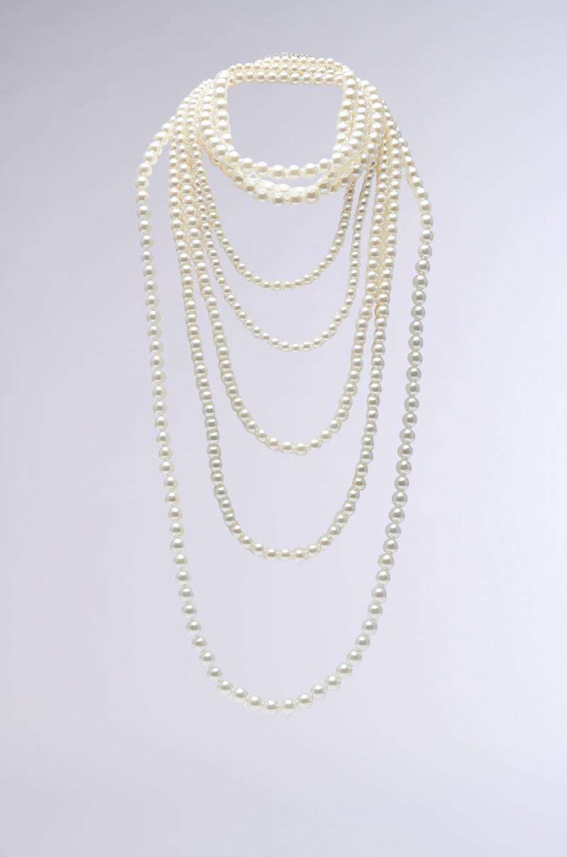 The PENELOPE STATEMENT PEARL NECKLACE features a long, elegant design with multiple strands that drape gracefully against a plain, light-gray background. The lustrous pearls exude a classic and sophisticated charm reminiscent of choker-style elegance.