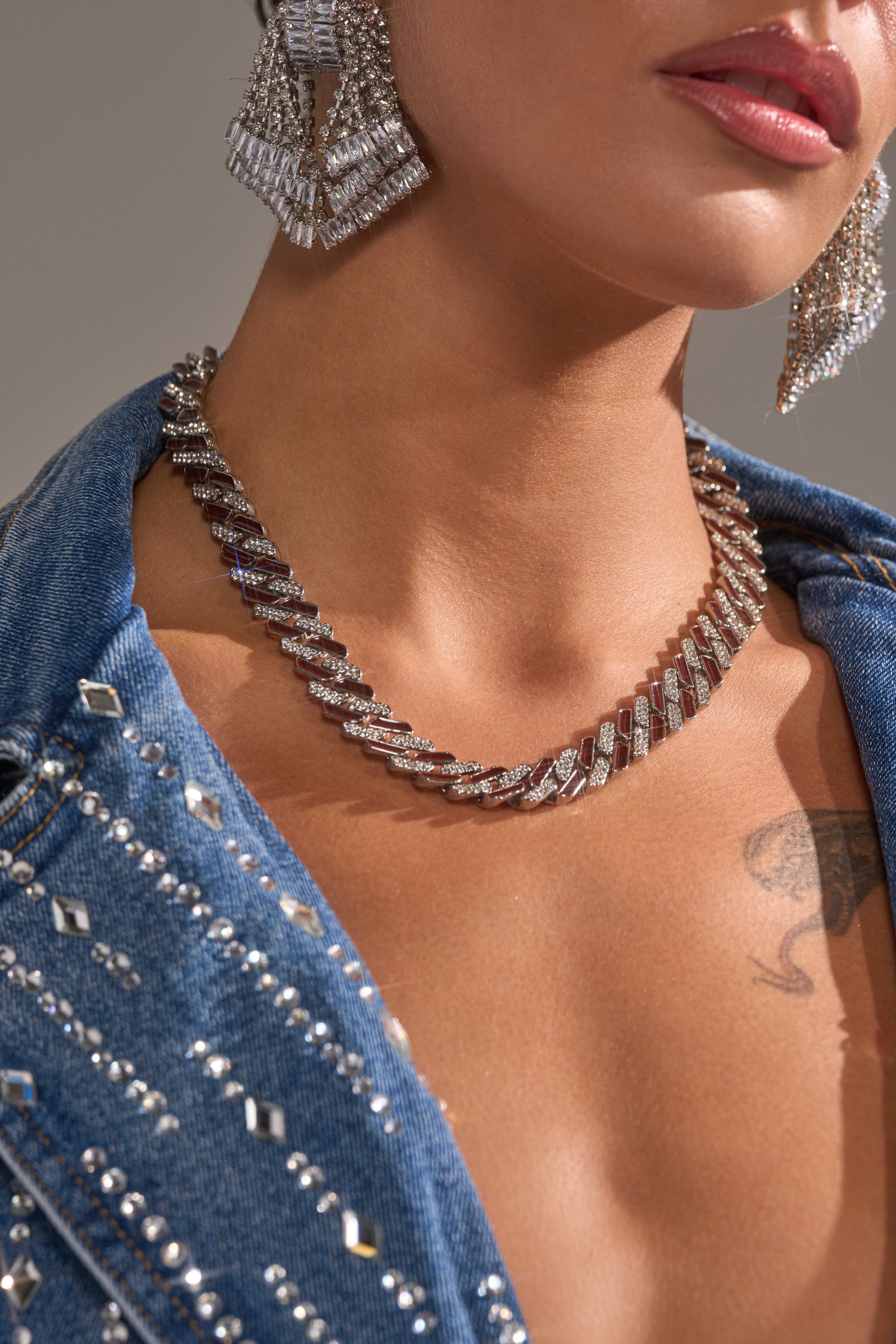 A woman wearing the CANDY CANE NECKLACE and matching earrings with a blue denim jacket featuring metallic studs. Only her lower face, neck, and upper chest are shown, revealing a tattoo near her collarbone. 1