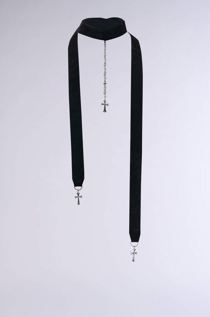 The CROSS MY HEART STATEMENT NECKLACE adorns the neck with a bold choker silhouette, showcasing two extended black velvet straps, each culminating in silver cross pendants. These straps gracefully hang in symmetry beside a delicate center cross, creating an elegant impression.