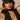 A person poses in a studded black outfit and a black cap with chain detailing, their long dark hair cascading as their hands rest behind their head. The CROSS MY HEART STATEMENT NECKLACE, featuring a cross pendant, elegantly drapes down their front.