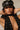 A person poses in a studded black outfit and a black cap with chain detailing, their long dark hair cascading as their hands rest behind their head. The CROSS MY HEART STATEMENT NECKLACE, featuring a cross pendant, elegantly drapes down their front.