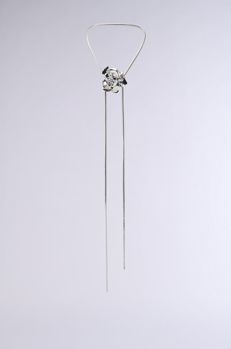The ASH ADJUSTABLE NECKLACE boasts a minimalist design with a silver wire frame, featuring a geometric centerpiece and two long dangling rods that resemble a metallic snake chain, all set against a plain light gray background.