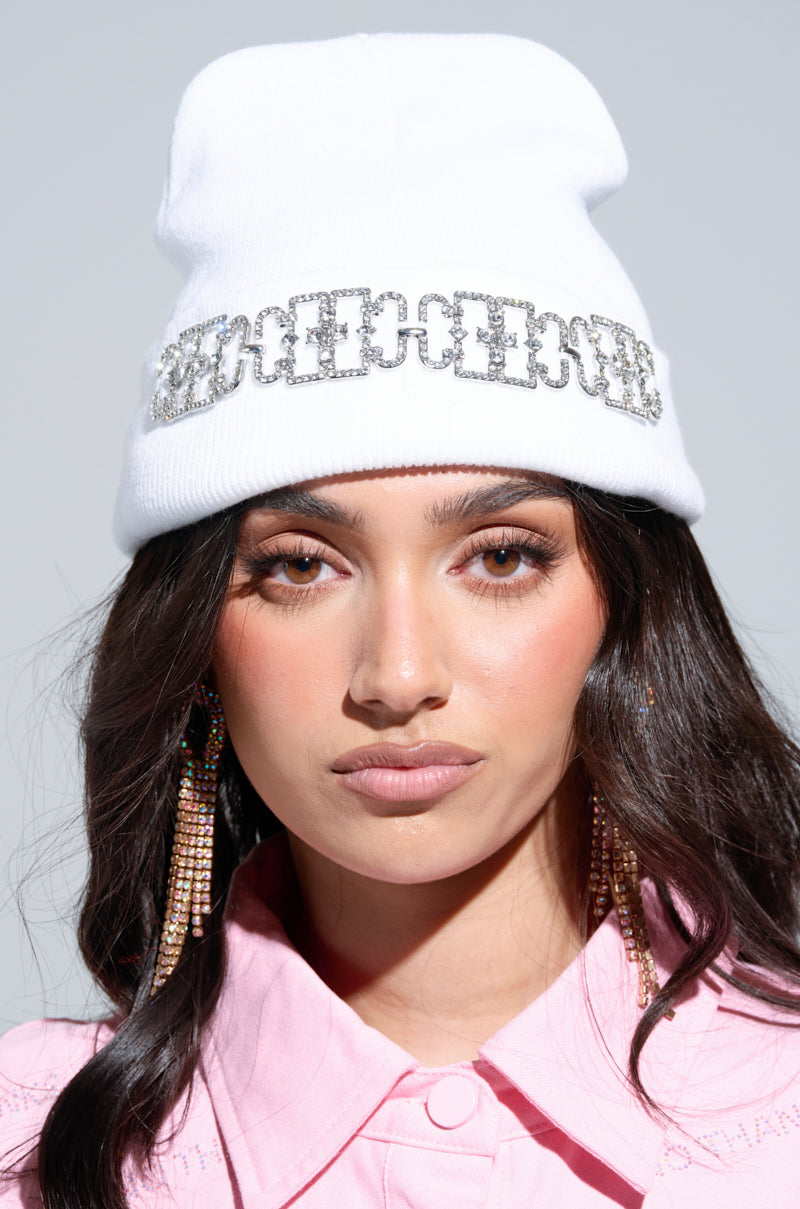 A person wearing a KIKI Rhinestone Beanie adorned with a chain design and paired with long, sparkling earrings. Their dark hair complements the pink collared shirt they are dressed in, while the neutral background highlights their striking accessories.