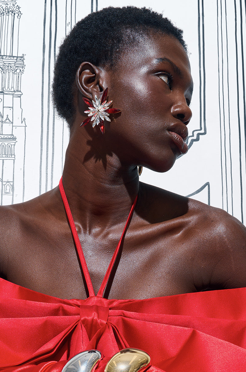 A person with short hair gazes to the side, showcasing a red strapless dress and the MAIN CHARACTER EARRING, which features large flower shapes with faux gold metallic accents. The background displays black and white line art of buildings.