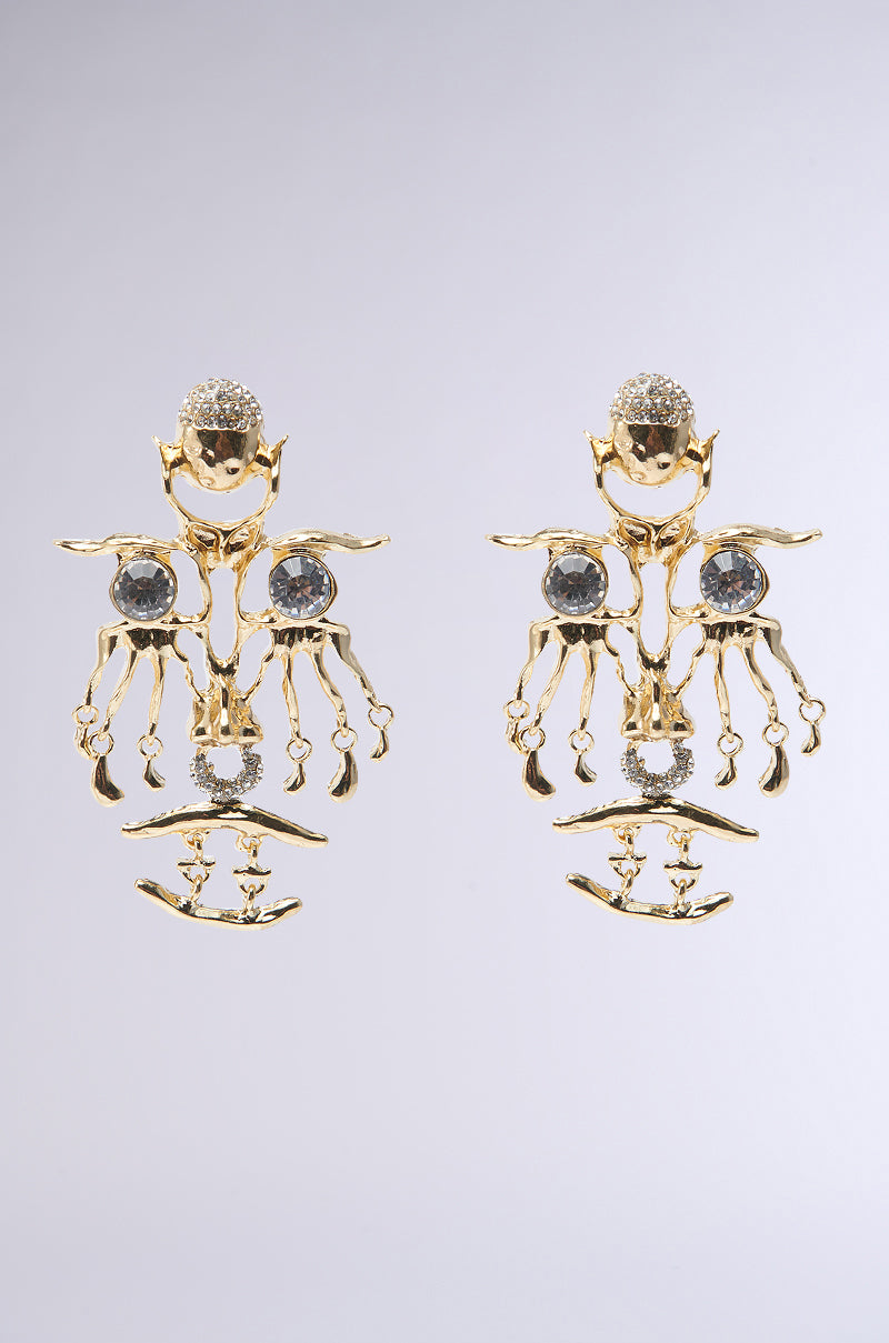 The OFF THE RUNWAY EARRING boasts rhinestone embellishments and intricate dangling elements, showcasing faux gold earrings shaped like abstract figures against a plain background.