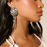 Profiled against soft lighting, a person with long dark hair showcases the TREAT ME EARRING—a large, ornate faux silver design with dangling elements. Complemented by subtle makeup, they're dressed in an elegant white lace garment that accentuates their smooth skin.