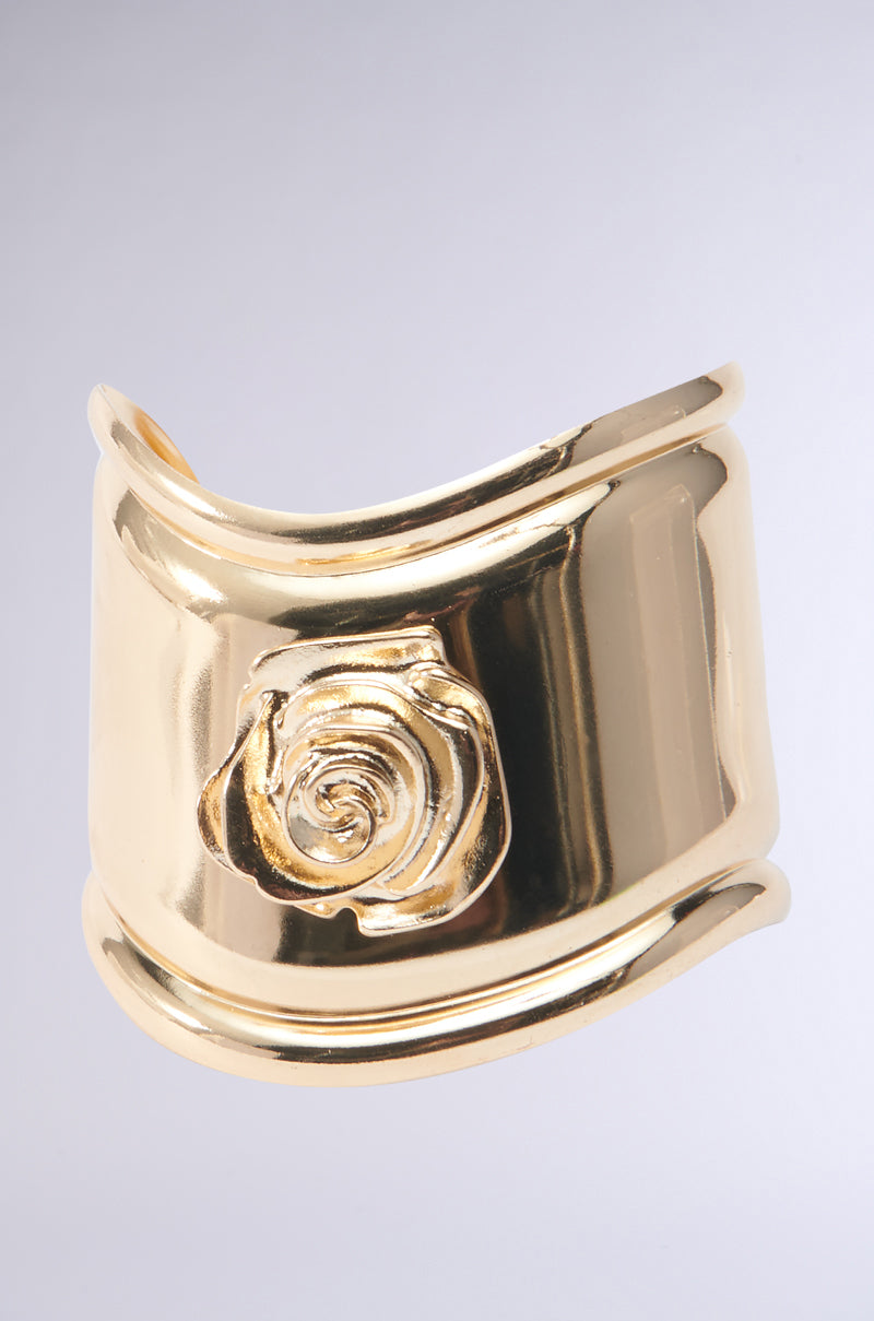 The SEAL WITH A ROSE BRACELET CUFF is a faux gold cuff bracelet that features a shiny finish and a raised rose design in the center. The bracelet boasts a smooth, polished surface with a slightly curved shape, set against a simple light gray background.