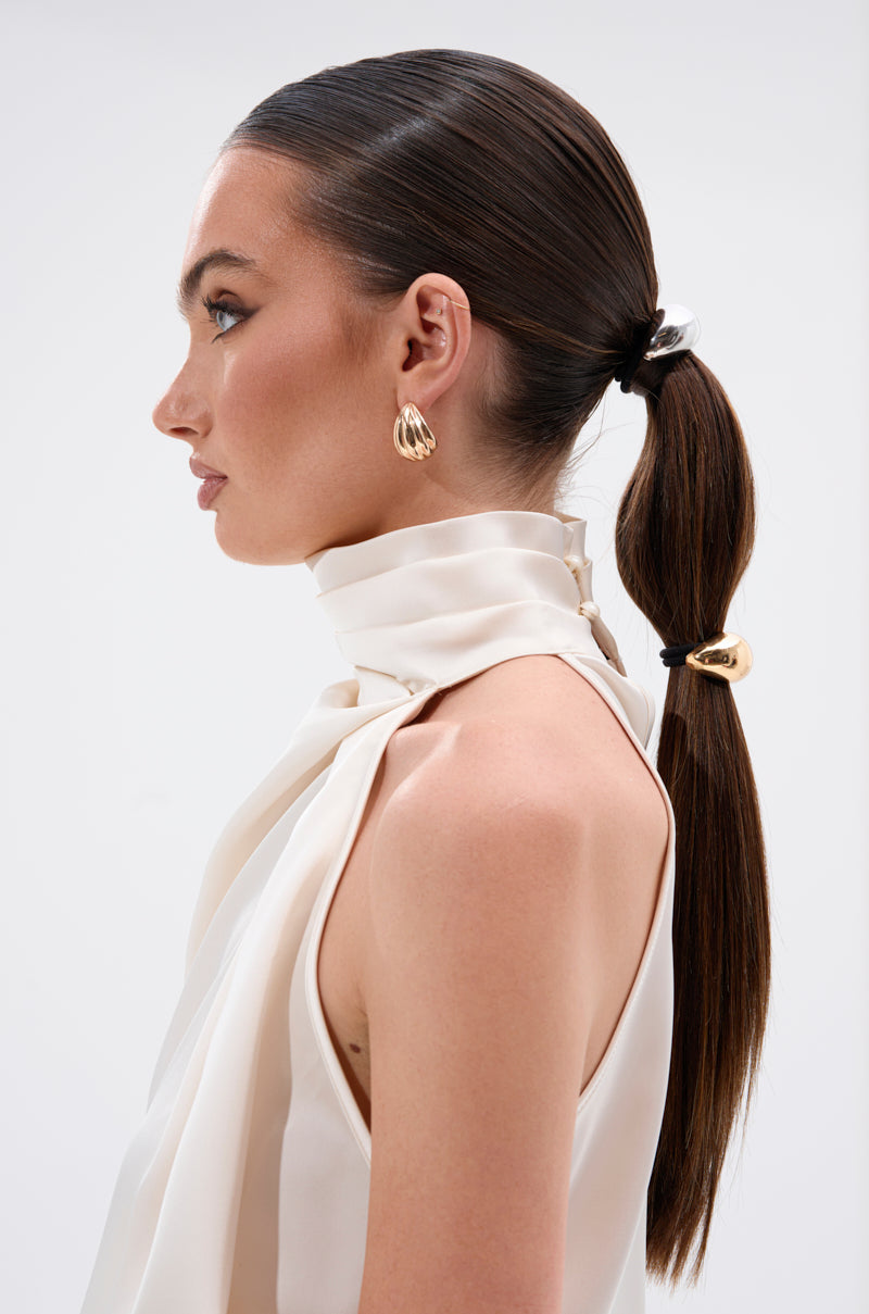 A woman in profile with straight dark hair styled in a segmented low ponytail, secured with BEST OF BOTH WORLDS HAIR TIES. She wears a sleeveless, high-neck white blouse and gold hoops against a plain backdrop.