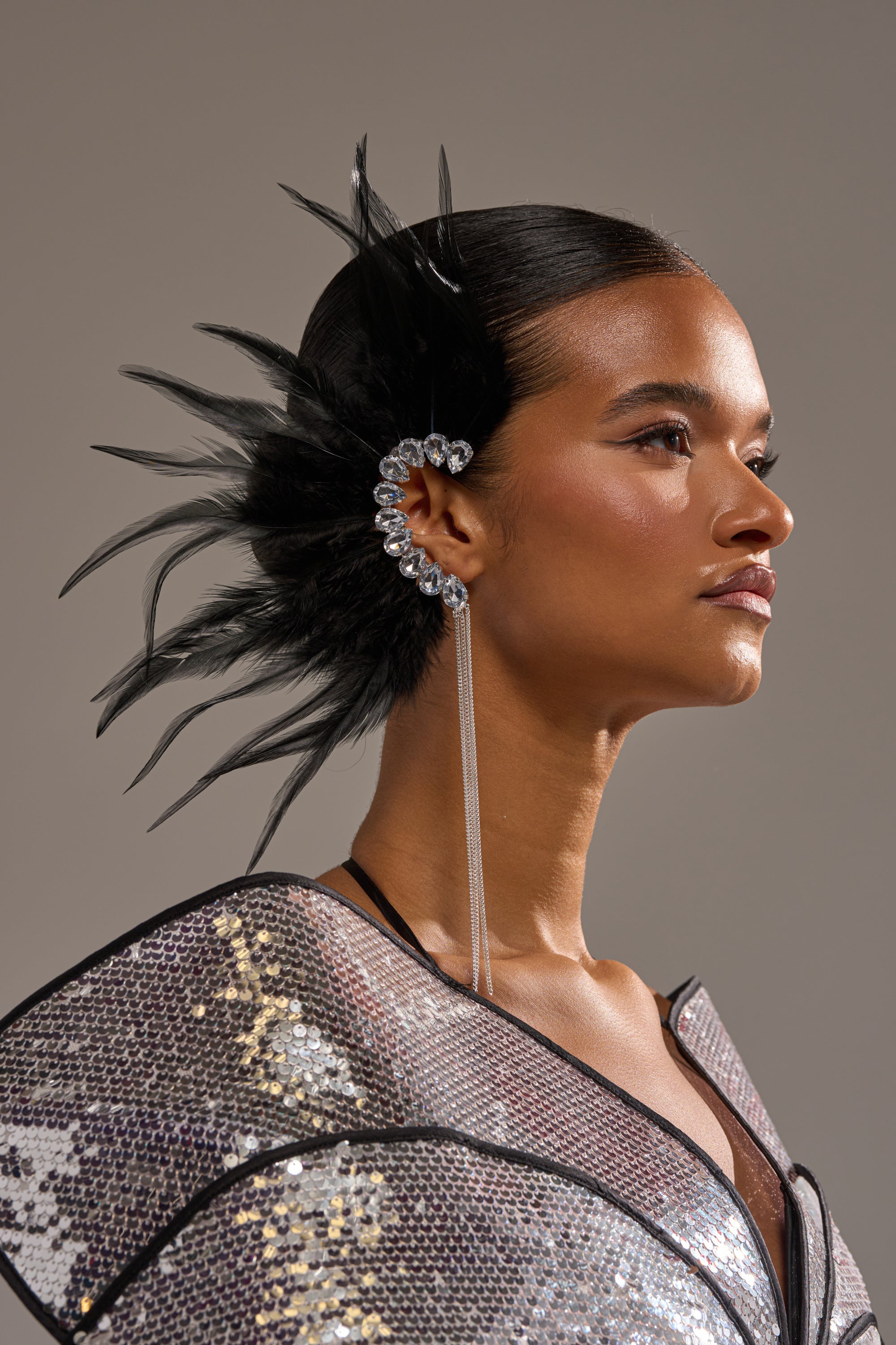 A woman with sleek, pulled-back hair wears the GLAM BIRD BLACK FEATHER EAR CUFF IN BLACK SILVER, featuring black feathers and crystals. Dressed in a metallic, structured-shoulder outfit, she gazes confidently to the right. 1