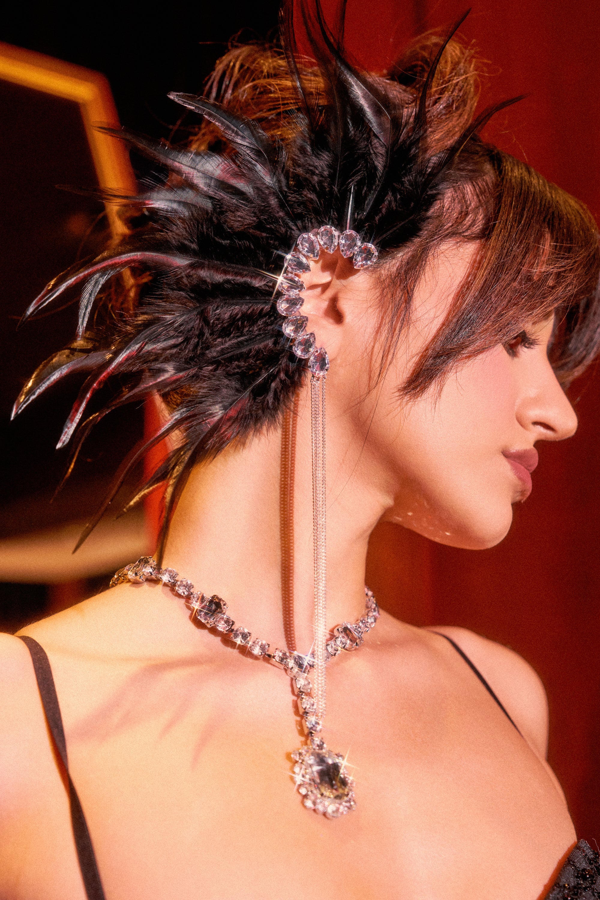 A woman with short, dark, spiky feathered hair wears the GLAM BIRD BLACK FEATHER EAR CUFF IN BLACK SILVER, a sparkling rhinestone ear cuff with a matching long chain earring and jeweled necklace, set against a red background. 0