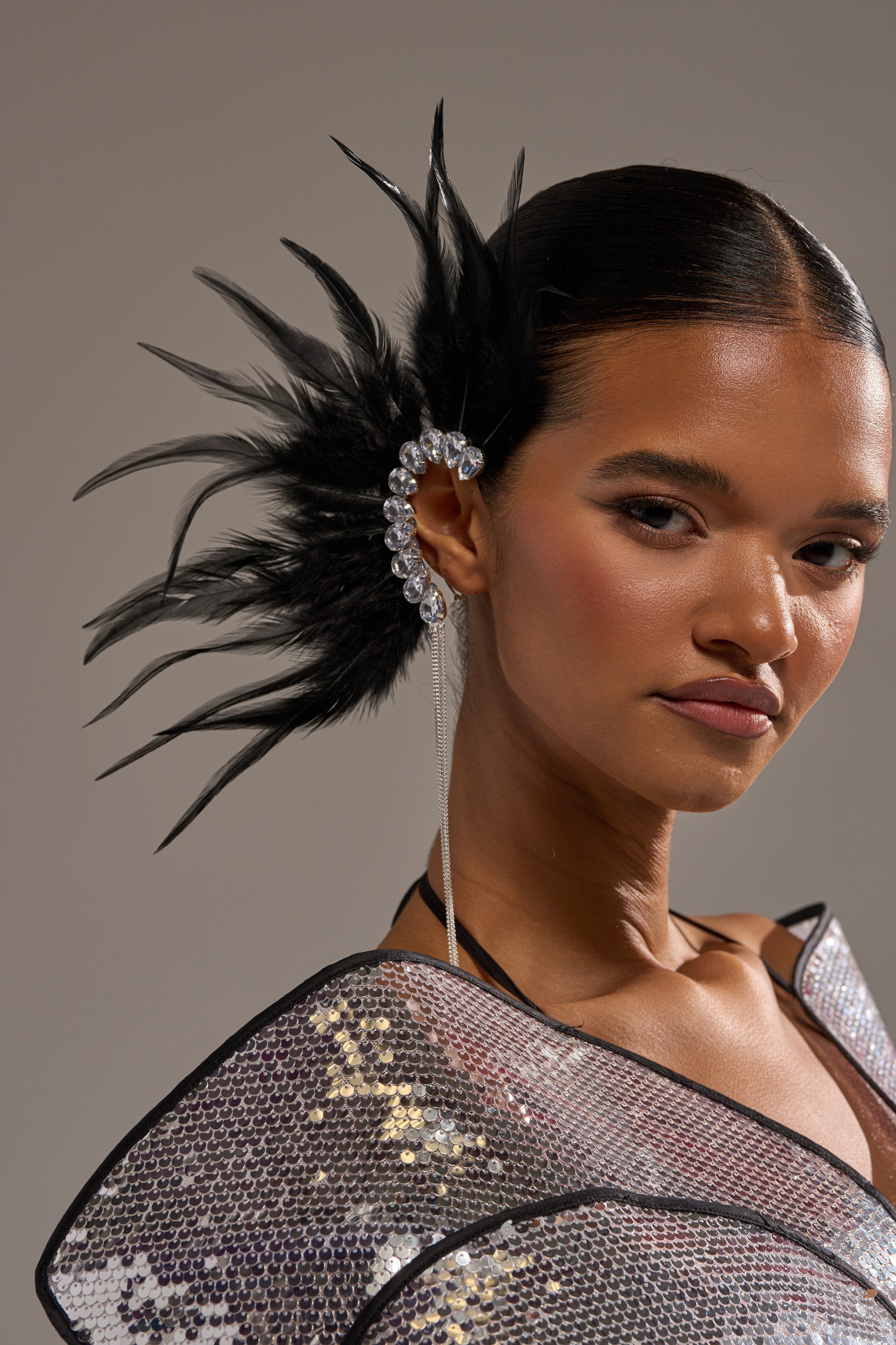 A woman with sleek hair wears the GLAM BIRD BLACK FEATHER EAR CUFF IN BLACK SILVER, featuring black feathers and crystal accents, paired with a shimmering sequin outfit as she confidently faces the camera. 2