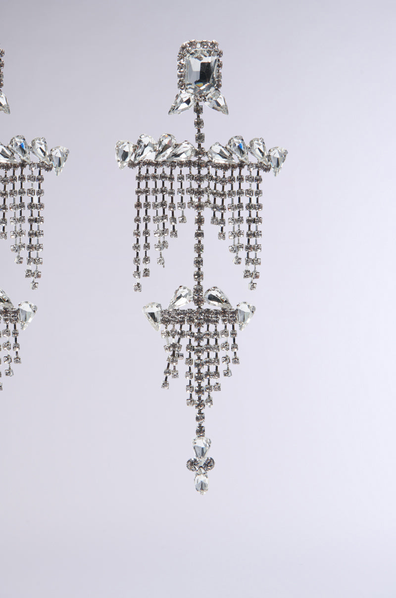 Close-up of the All Night Long Statement Earring. This ornate chandelier earring showcases teardrop-shaped jewels, arranged in a geometric pattern with sparkling crystals forming cascading tiers of dangling strands, set against a neutral background.