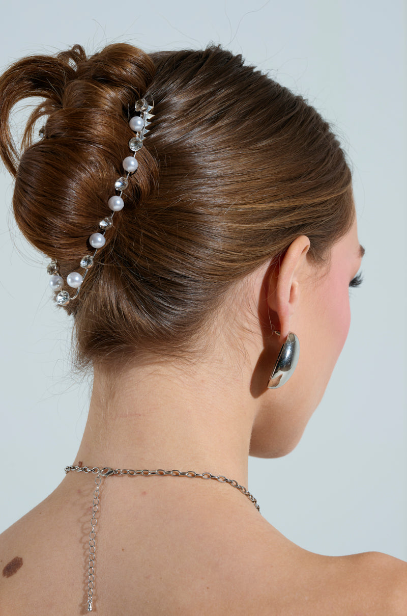 A woman with brown hair styled in a bun wears the REGAL RHINESTONE HAIR CLIP. She dons metallic silver earrings and a chain necklace draping down her back against a light background, with her face turned away from the camera, emphasizing the subtle elegance of her accessories.