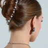 A woman with brown hair styled in a bun wears the REGAL RHINESTONE HAIR CLIP. She dons metallic silver earrings and a chain necklace draping down her back against a light background, with her face turned away from the camera, emphasizing the subtle elegance of her accessories.