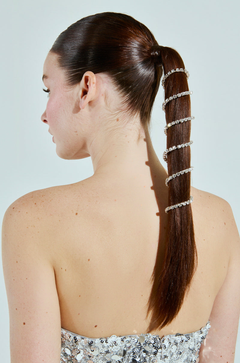 A woman with smooth, dark brown hair styled in a high ponytail showcases the NEVER SPIRALING BLING PONY WRAP, adorned with silver beads. Her sequined strapless top enhances the side-back view captured against a light background.
