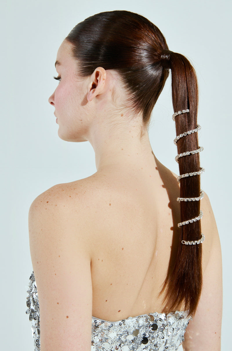 A woman in a strapless silver sequin dress with a sleek brown ponytail stands side-on. Her hair shimmers with rhinestone embellishments from the NEVER SPIRALING BLING PONY WRAP, adding sparkling rings down its length against the plain backdrop for a glamorous touch.