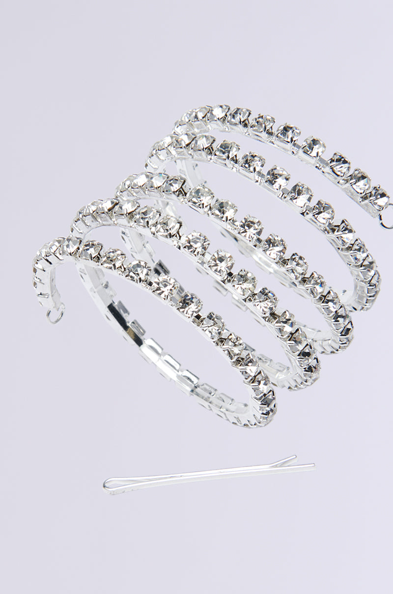 The NEVER SPIRALING BLING PONY WRAP, a silver spiral bracelet with rhinestone embellishments, elegantly floats against a light gray background. Beneath it, a delicate silver hairpin adds an exquisite touch of style.