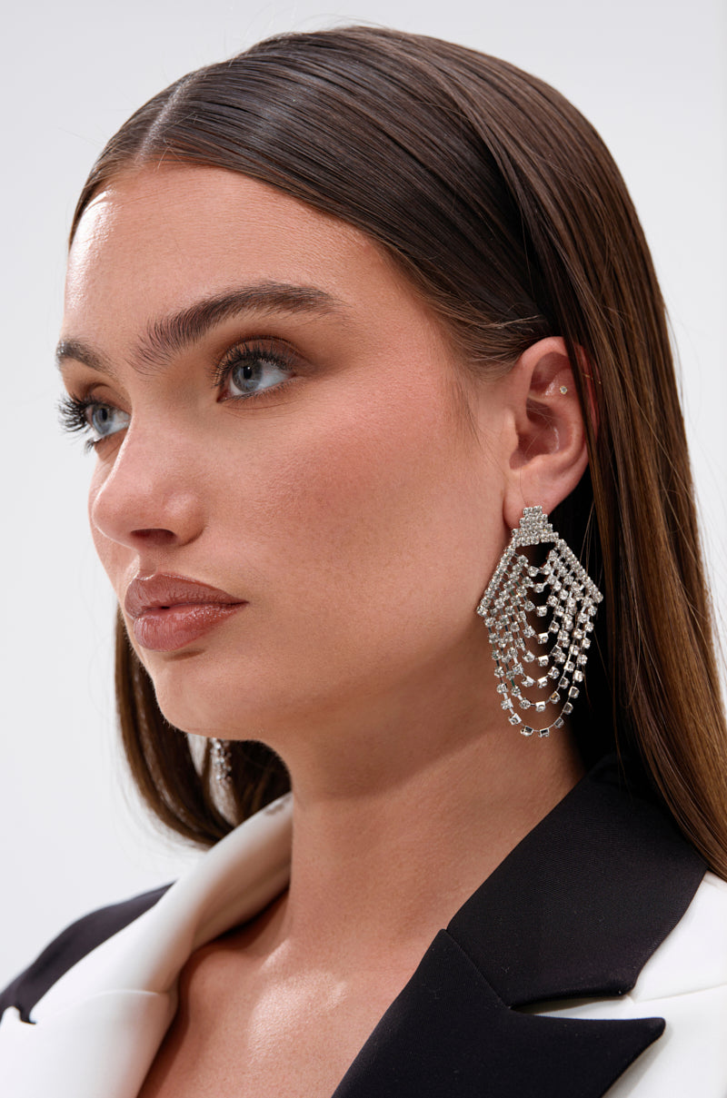 A woman with straight brown hair and blue eyes wears a white and black blazer and the TO THE GALA EARRING IN SILVER from AKIRA Label—large chandelier-style earrings adorned with sparkling rhinestones. She gazes slightly to the side.