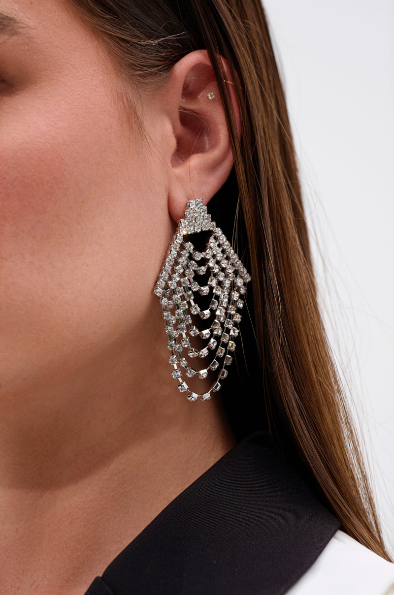 Close-up of a person wearing the AKIRA Label TO THE GALA EARRING IN SILVER—a large, chandelier-style accessory adorned with sparkling rhinestones. Their straight brown hair is tucked behind the ear, paired with a black and white top.