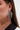 Close-up of a person wearing the AKIRA Label TO THE GALA EARRING IN SILVER—a large, chandelier-style accessory adorned with sparkling rhinestones. Their straight brown hair is tucked behind the ear, paired with a black and white top.