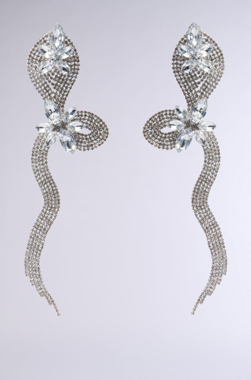 The TILLY STATEMENT EARRING boasts a sophisticated allure with its elongated, shimmering earrings showcasing intricate silver and crystal details. Featuring a floral motif and cascading strands, it’s complemented by a snake-inspired lobe post beautifully embellished with delicate rhinestones set against a minimal backdrop.