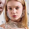Two models wearing sparkling crystal earrings and necklaces. The main focus is on one model with long blonde hair and large TAYTAY STATEMENT EARRINGS that feature a swooping abstract design, framed between silver chains. The background is softly out of focus with a decorative pattern.