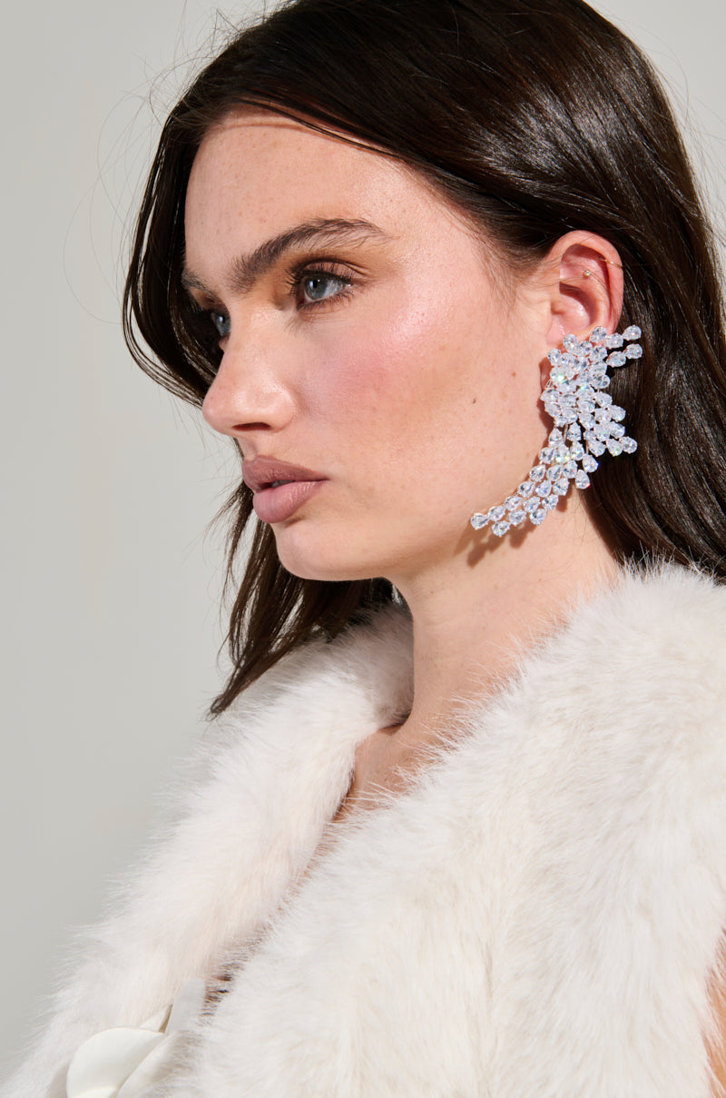 A woman with long brown hair is adorned with the TAYTAY STATEMENT EARRING, an intricate crystal ear cuff shaped like a cluster of leaves. With a soft, neutral expression, she wears a white fur-like vest over a silky white top, set against a light background. 2
