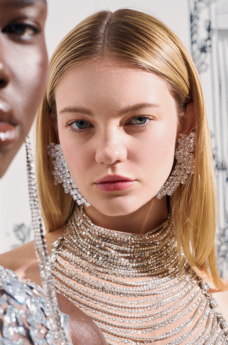 Two models wearing sparkling crystal earrings and necklaces. The main focus is on one model with long blonde hair and large TAYTAY STATEMENT EARRINGS that feature a swooping abstract design, framed between silver chains. The background is softly out of focus with a decorative pattern. 0