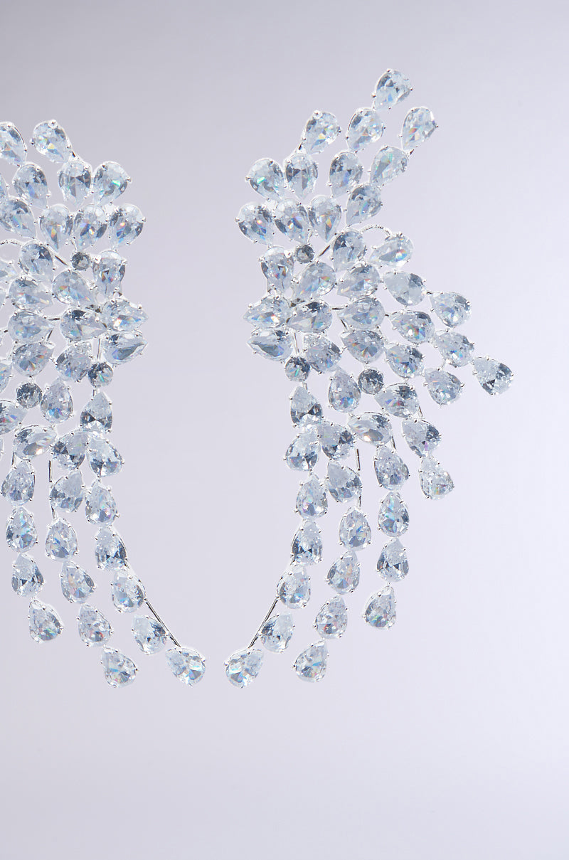 The TAYTAY STATEMENT EARRING showcases an intricate and delicate swooping abstract design in silver, embellished with numerous clear teardrop rhinestones arranged in a cascading pattern against a smooth gray background, creating a stunning and sparkling effect. 4