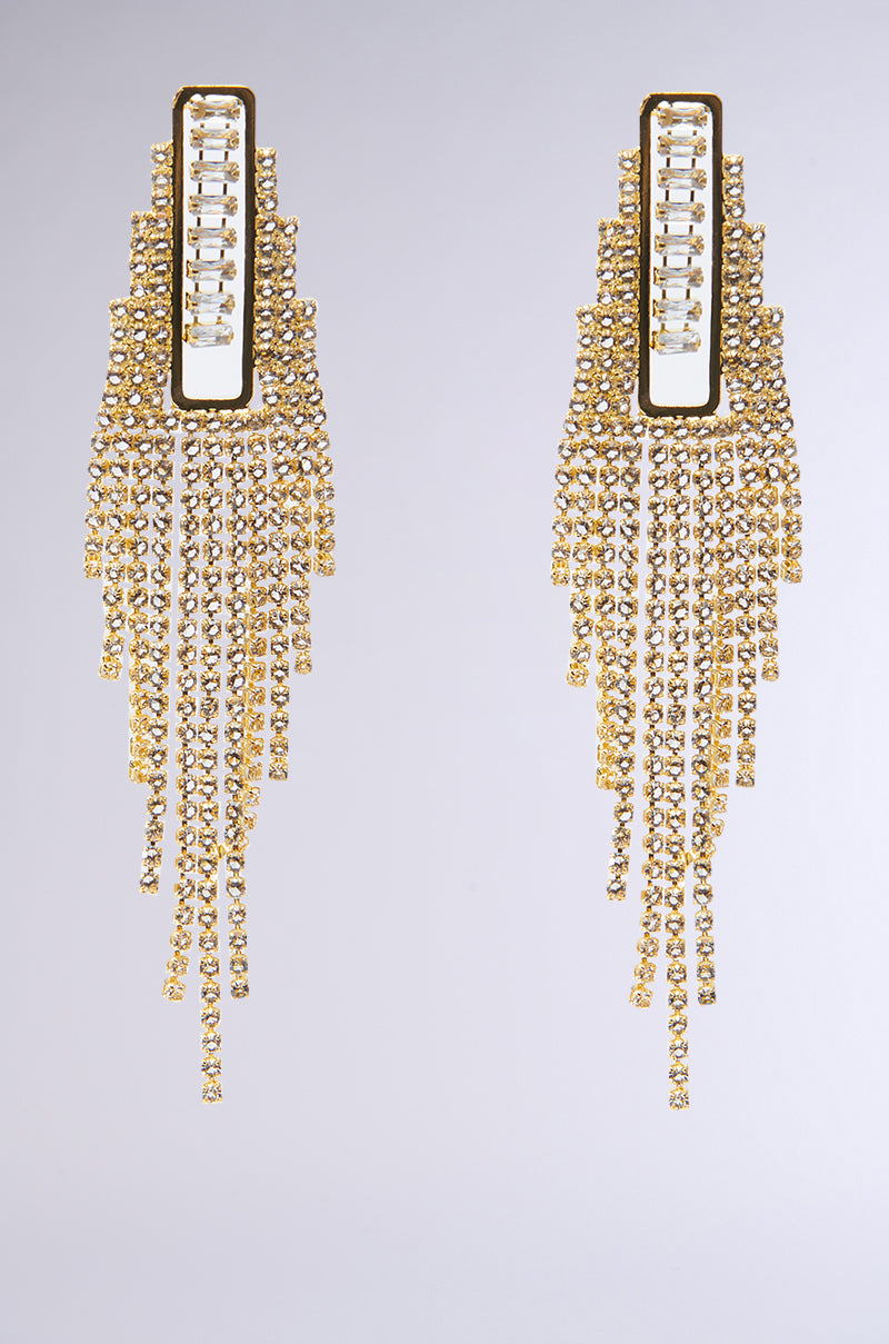 The ITS GIVING SEASON EARRING IN GOLD features a pair of long, gold-toned earrings with elegant rhinestone fringe and cascading strands of sparkling crystals, creating a chandelier effect against a plain background.