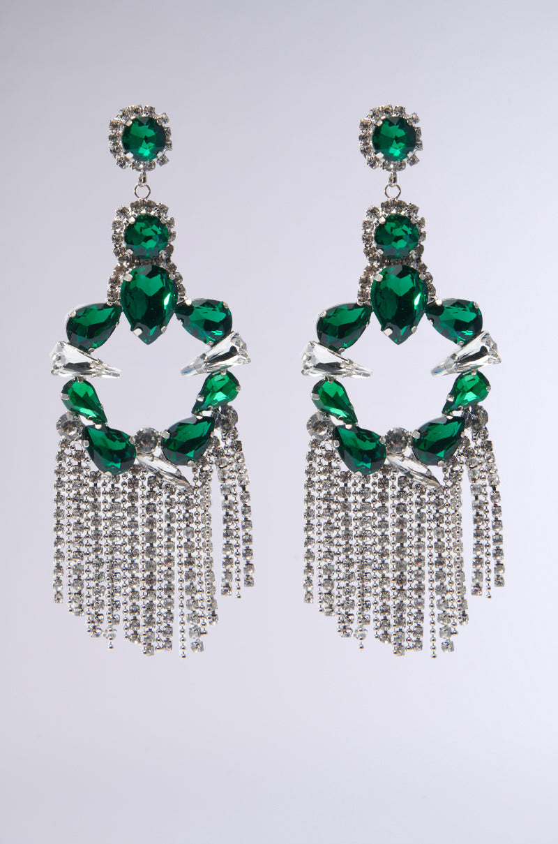Introducing the EMERALD DREAMS STATEMENT EARRING: these striking earrings showcase large green gemstones in a circular lobe post design, accentuated by silver details. Each earring is adorned with multiple strands of rhinestone fringe, offering a spectacular display against its light gray backdrop. 0