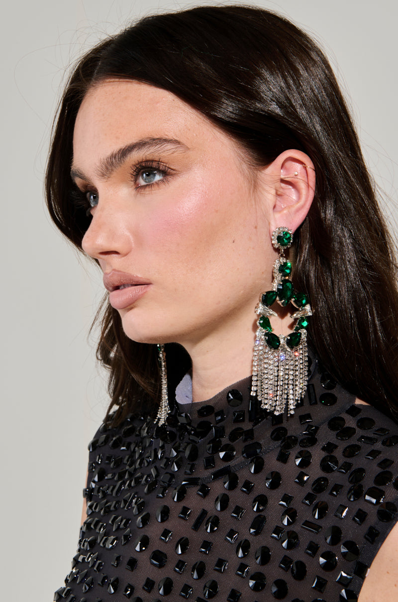 A woman with long brown hair wears striking Emerald Dreams Statement Earrings and a black top adorned with sparkling embellishments. She gazes to the side against a neutral background, her statement earrings adding an elegant allure. 1