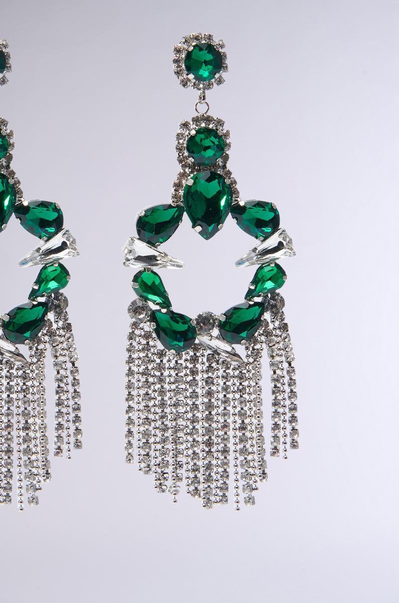 A close-up of the ornate Emerald Dreams Statement Earring showcases its large green gemstones and clear crystals. The earring is enhanced by hanging strands of smaller sparkling stones, creating a striking Rhinestone Fringe. The neutral background accentuates the jewelry’s intricate design. 3