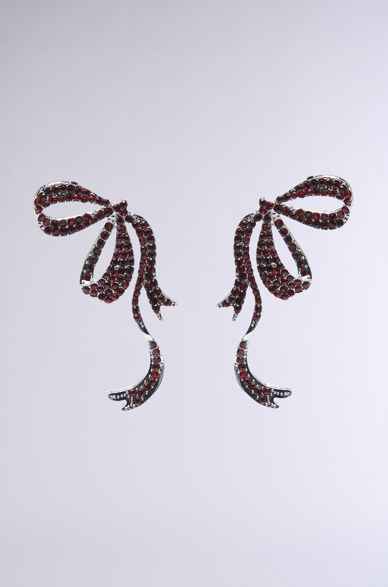 The FEMME BOW EARRING features a sleek metallic base with flowing ribbon-like loops, adorned with garnet-inspired rhinestones. Set against a plain gray background, the symmetrical design exudes timeless charm.