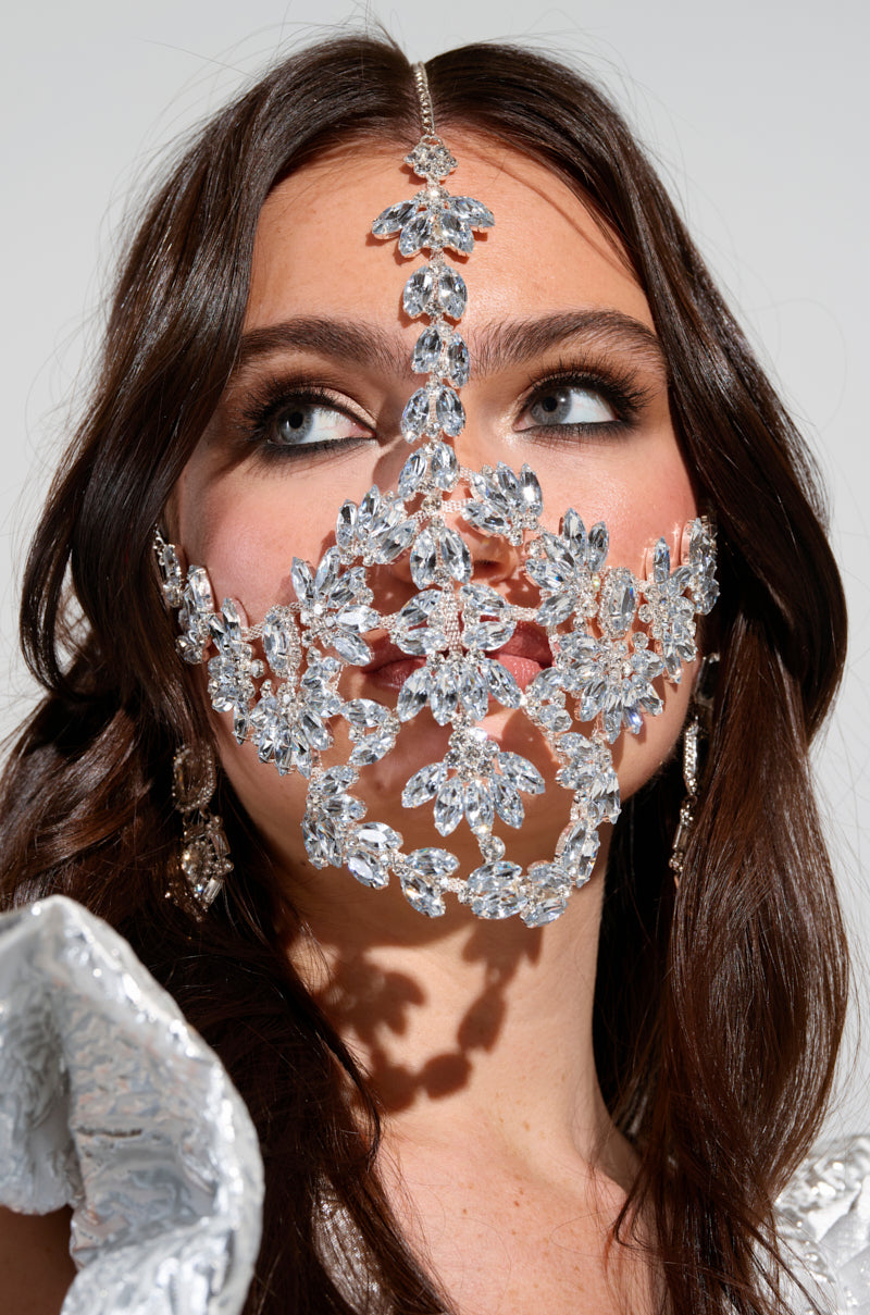 A woman with long brown hair wears an elaborate silver face mask featuring floral designs, sparkling crystals, and teardrop gemstone embellishments. As she looks upward, her nose and mouth are covered by the mask. Her white garment decorated with sequins is partially visible beneath the TAKE MY PIC STATEMENT HEADPIECE. 0