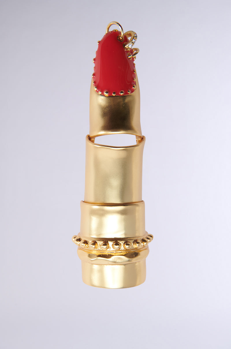 The NAILS DID BENDABLE RING features a faux gold sculptural design with three stacked segments resembling a finger, complete with a red-painted fingernail embellished with small gold studs and a sparkling ring at the base. It is set against a plain, light background. 0