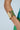 A close-up of a person’s arm wearing the GOLDEN GODDESS CUFF, a wide, textured metallic faux gold bracelet. The person is dressed in green and blue pleated fabric against a plain, light background.