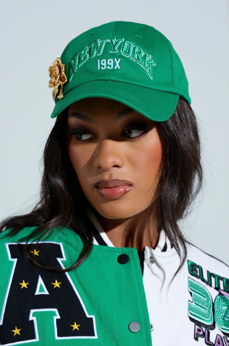 A woman in a "THIS IS MY CITY HAT" with an adjustable strap, "New York 199X" embroidery, and a gold rose pin. Her long dark hair contrasts her green and white jacket with large letter patches and a star pattern, as she gazes sideways against a plain background. 0