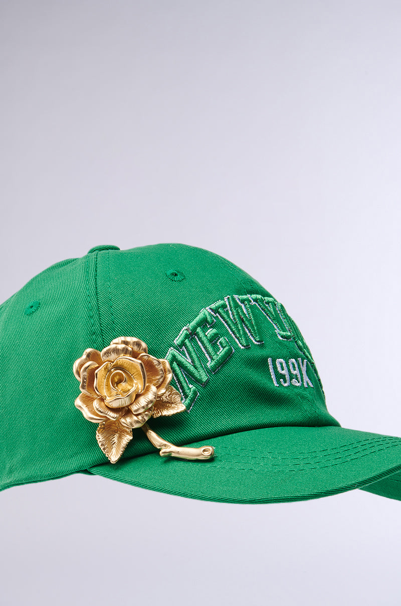 The "THIS IS MY CITY HAT" is a green cap with "NEW YORK 199X" in white, featuring an adjustable strap for comfort. Adorning the brim is a large gold rose embellishment on a plain gray backdrop. 4