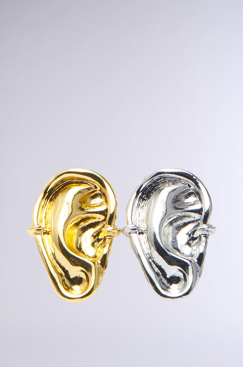 The HEARD YOU RING SET features two sculptural earrings resembling abstract faces, one in faux gold and the other in metallic silver, elegantly hanging as if sharing secrets against a plain light gray background.