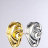 The HEARD YOU RING SET features two sculptural earrings resembling abstract faces, one in faux gold and the other in metallic silver, elegantly hanging as if sharing secrets against a plain light gray background.
