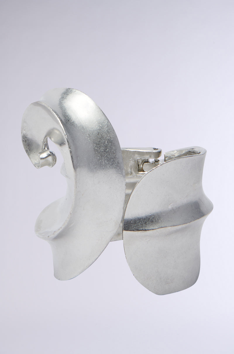 An abstract silver sculpture features interlocking curves and angles against a light gray background. Reminiscent of a modern accessory like the KANDYCE BRACELET, it has an artistic design with a smooth metallic finish.