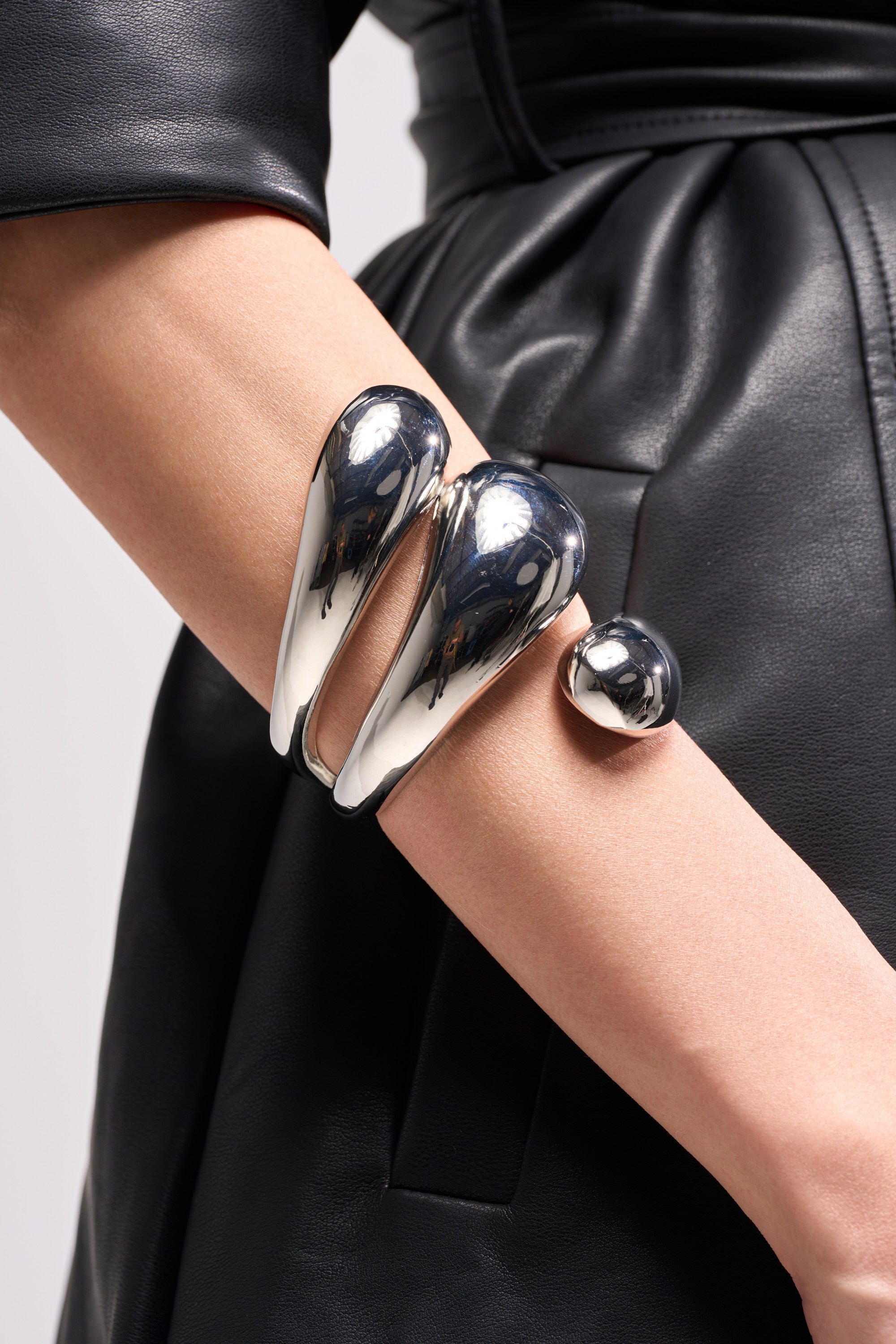 Close-up of a person wearing the shiny, chunky LOVER BRACELET CUFF on their forearm. Paired with a black belted leather outfit, the cuff features smooth, rounded forms and a modern, artistic design. 3