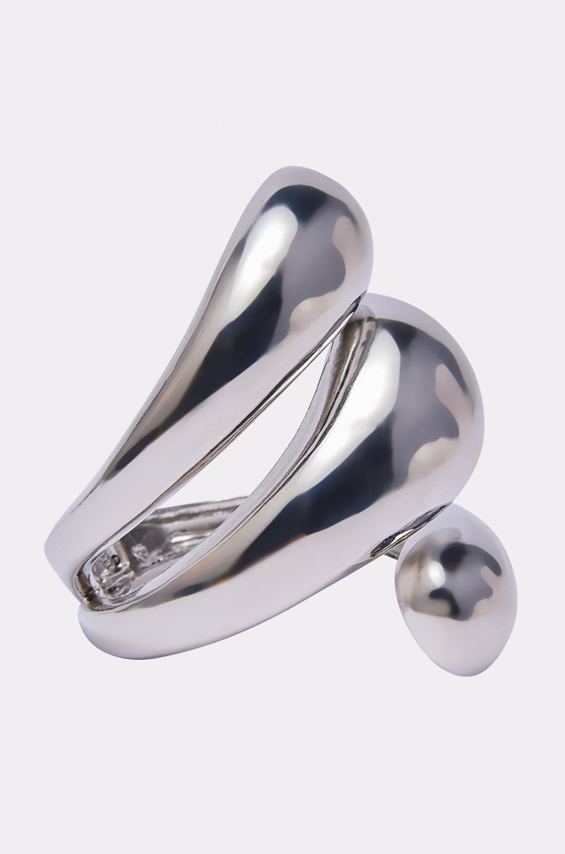 The LOVER BRACELET CUFF is a shiny, modern silver cuff with a bold, sculptural wave-inspired design, featuring smooth curved bands and a large round accent, displayed against a simple light background. 2