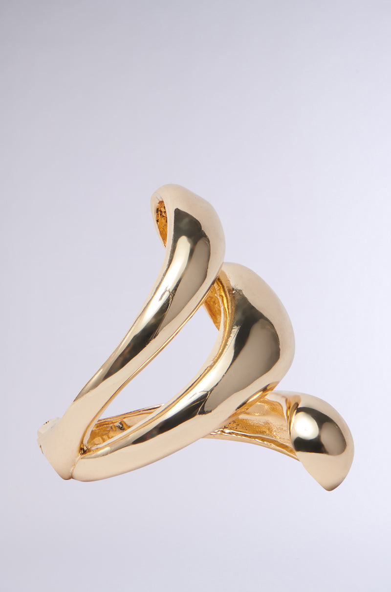 The IT IS WHAT IT IS BRACELET CUFF is a modern, abstract faux gold piece featuring three intertwined smooth loops in a sculptural design, reminiscent of luxury jewelry, set against a plain gray background. 3