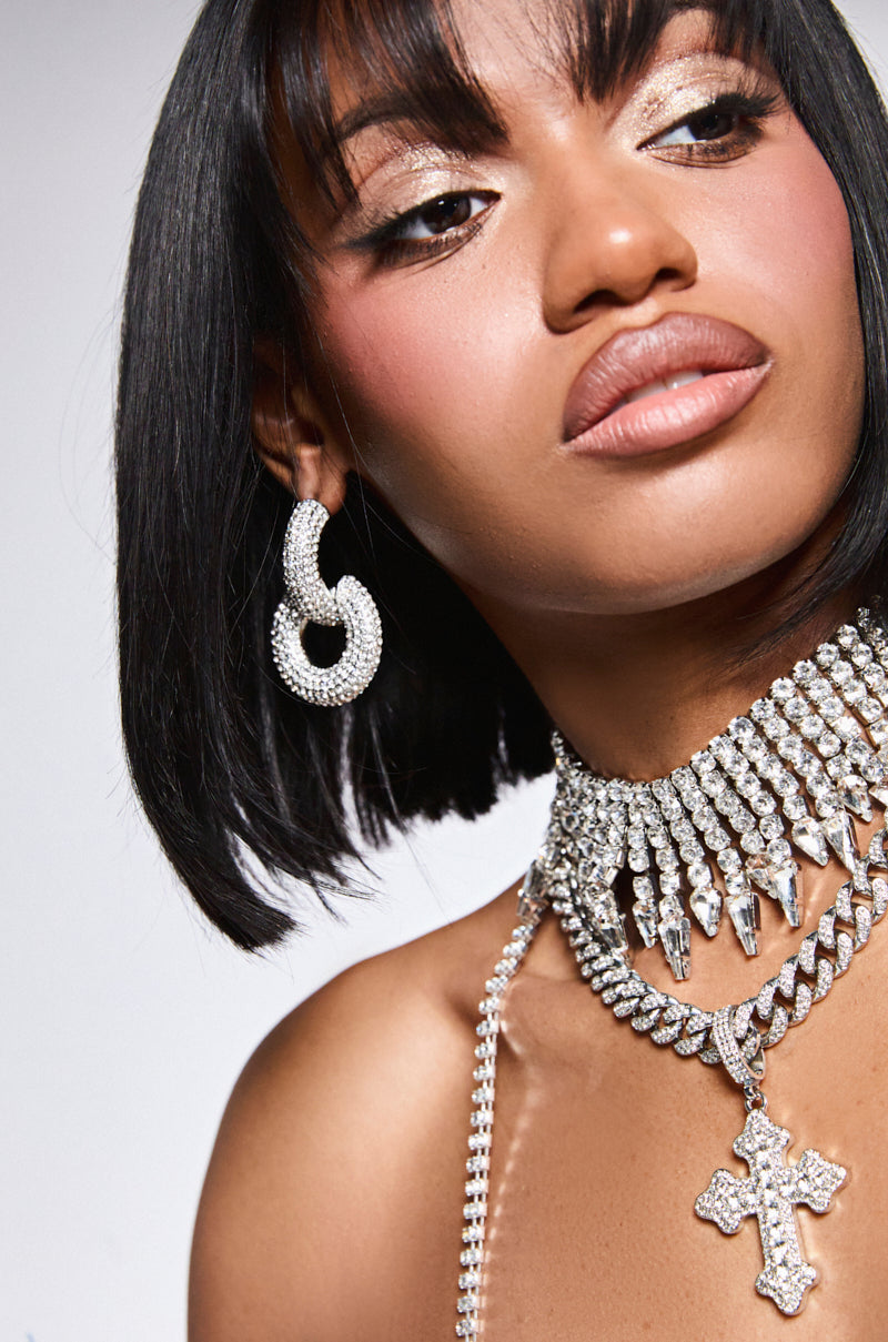 Against a simple background, a person with black hair and shimmering makeup wears an elegant AKIRA Label "IM ON IT EARRING," featuring interlocking hoops encrusted with sparkling rhinestones, along with a necklace adorned with large pointed crystals and a cross pendant.
