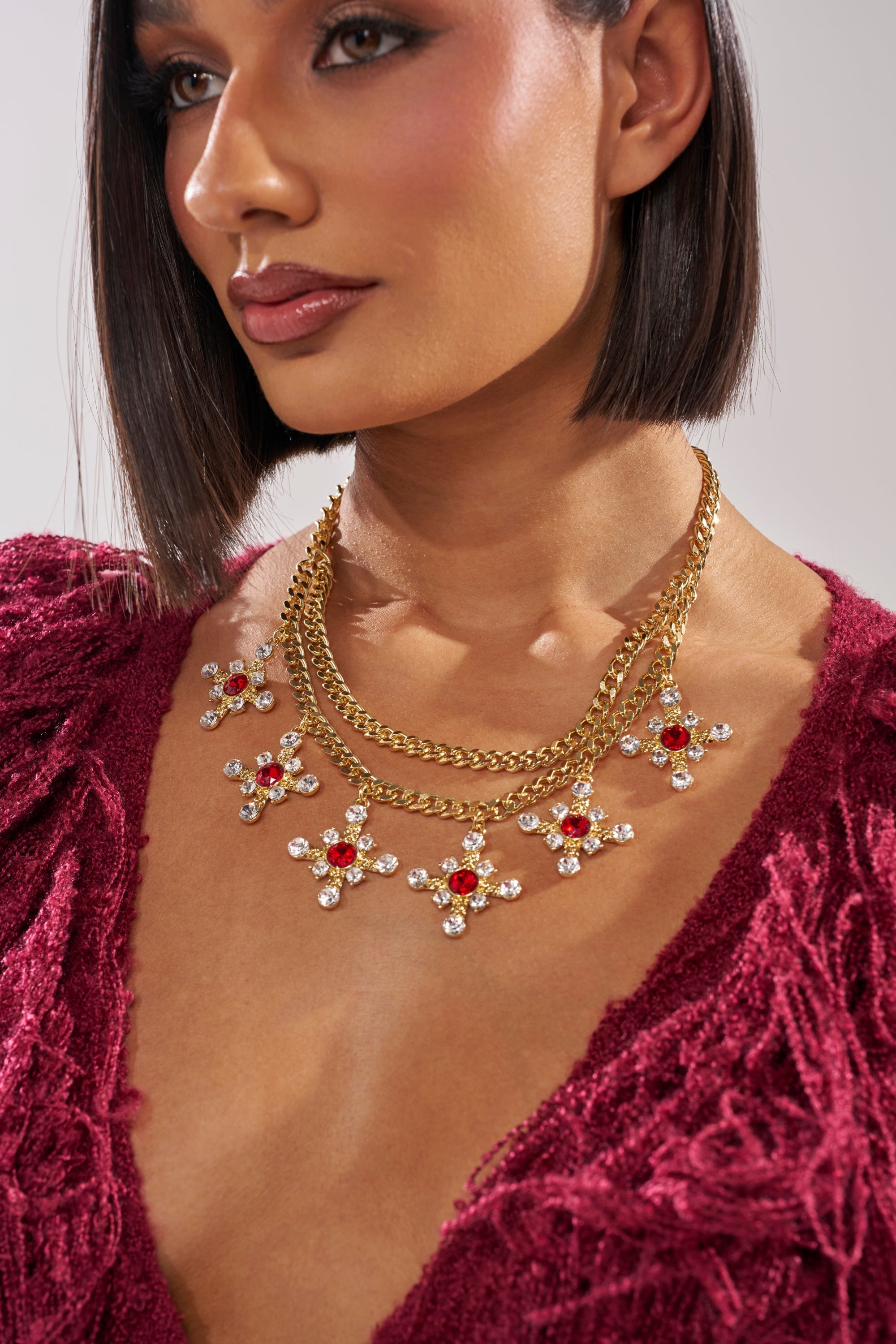 A woman wears a deep red textured top and the MAXIMUM GLAM NECKLACE, featuring gold layers with red and clear gemstone floral pendants. Cross crystal charms accent her sleek chin-length hair for a modern, elegant look. 1